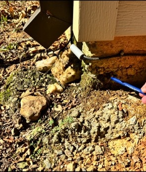 Gutter downspout discharging directly against a house foundation corner, with soil washout and heavy moss on the concrete.