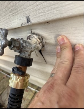 Exterior hose bib with the vinyl siding panel split open around the mounting flange, leaving a gap into the wall cavity.
